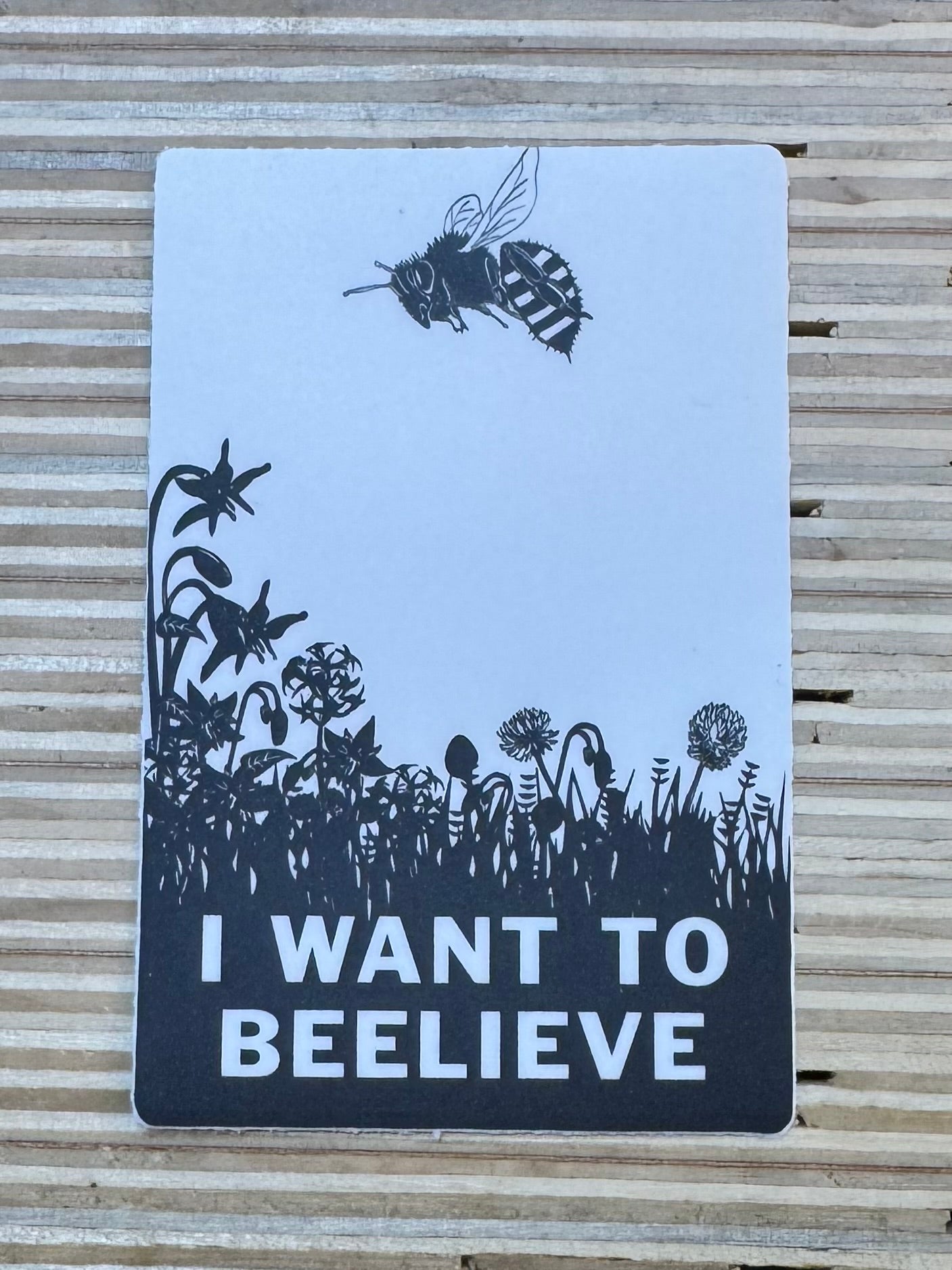 Sticker with black illustrations of a Bee, flowers, and a bee on a light blue background, with text 'I Want to Beelieve' on a wooden surface.