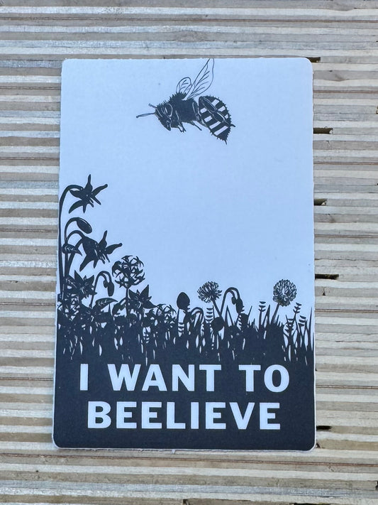 Sticker with black illustrations of a Bee, flowers, and a bee on a light blue background, with text 'I Want to Beelieve' on a wooden surface.